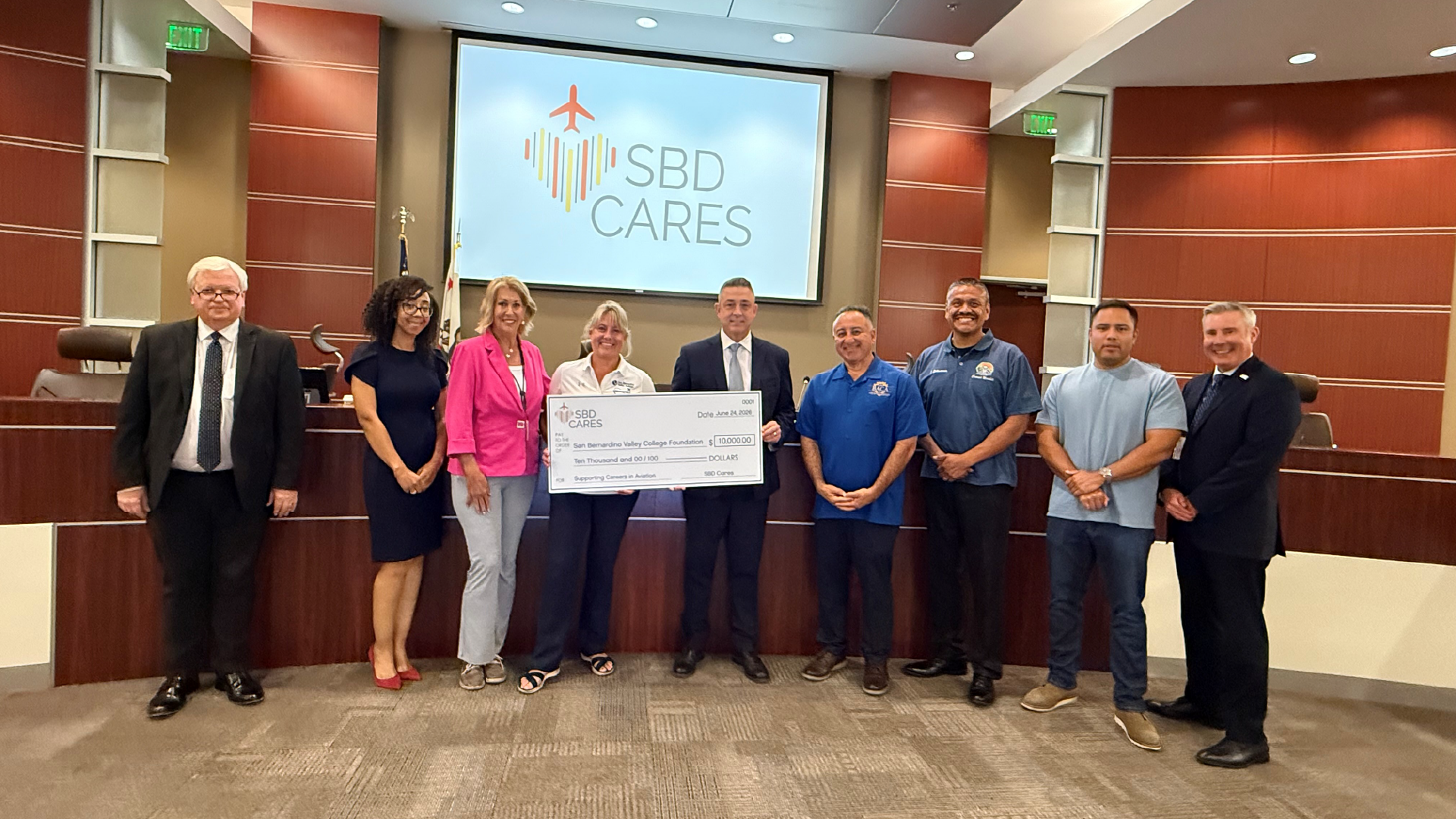 A group of nine people stand in a boardroom holding an oversized check under a screen that reads “SBD Cares.